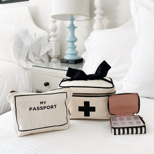 Bag-all First Aid Travel Gift Set 3-Pack in cream: cotton passport pouch, first aid box with black bow, and striped pill case displayed on white bedding with blue table lamp