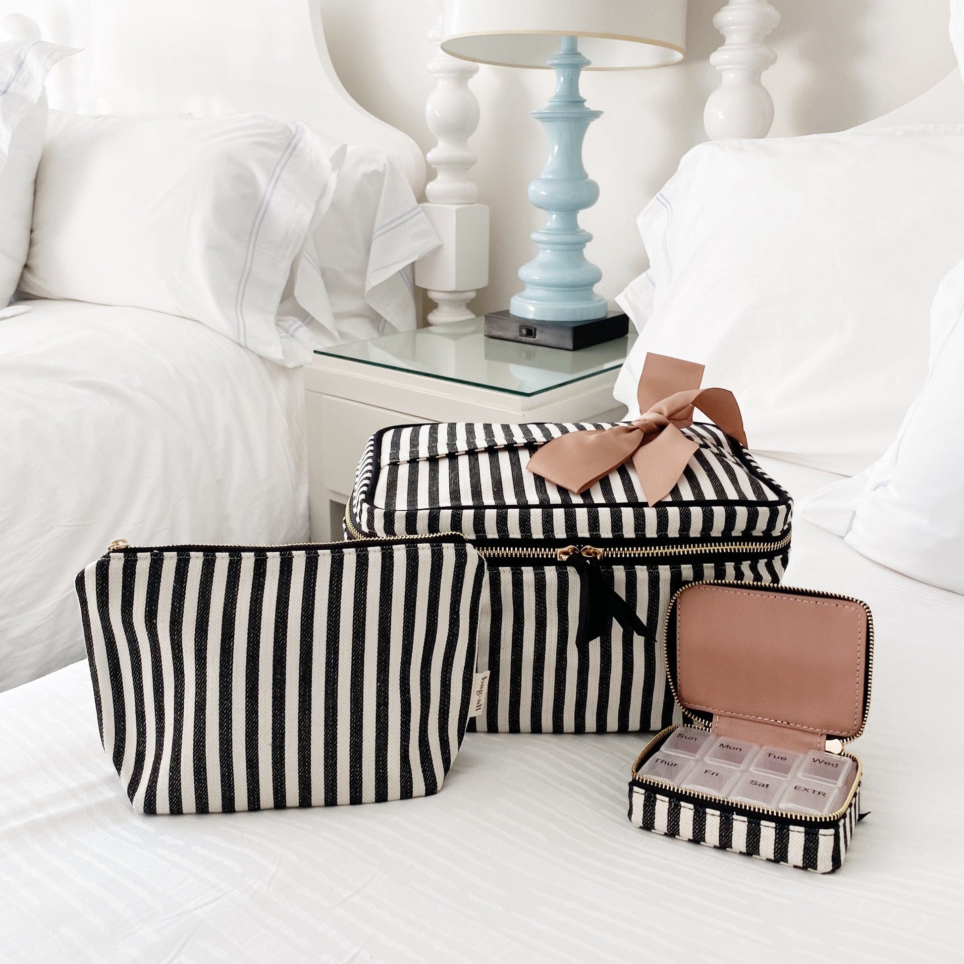 Bag-all Cutest Striped Travel Gift Set 3-Pack featuring black and white striped cotton makeup bag, toiletry case, and pill organizer with pink bow accent on crisp white bedding