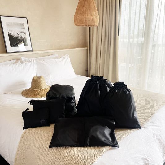 Bag-all Compression Cubes & Packing Bags Set displayed on white hotel bed with straw hat, featuring 8 black organizers in various sizes for efficient travel packing and organization