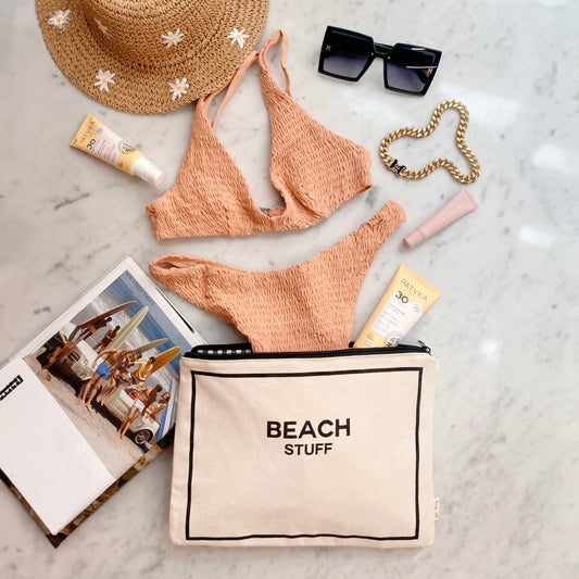 Bag-all Beach Pouch with text 'BEACH STUFF' displayed with peach bikini, straw hat, sunglasses, sunscreen, and travel essentials on marble surface. Waterproof cotton storage for beach day organization.