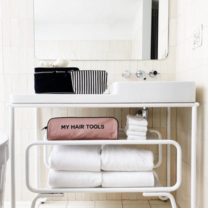 Bag-all Medium Box Makeup & Toiletry in Black shown in modern bathroom setting with striped lining and gold zipper, displayed alongside white towels and hair tools case on bathroom shelf