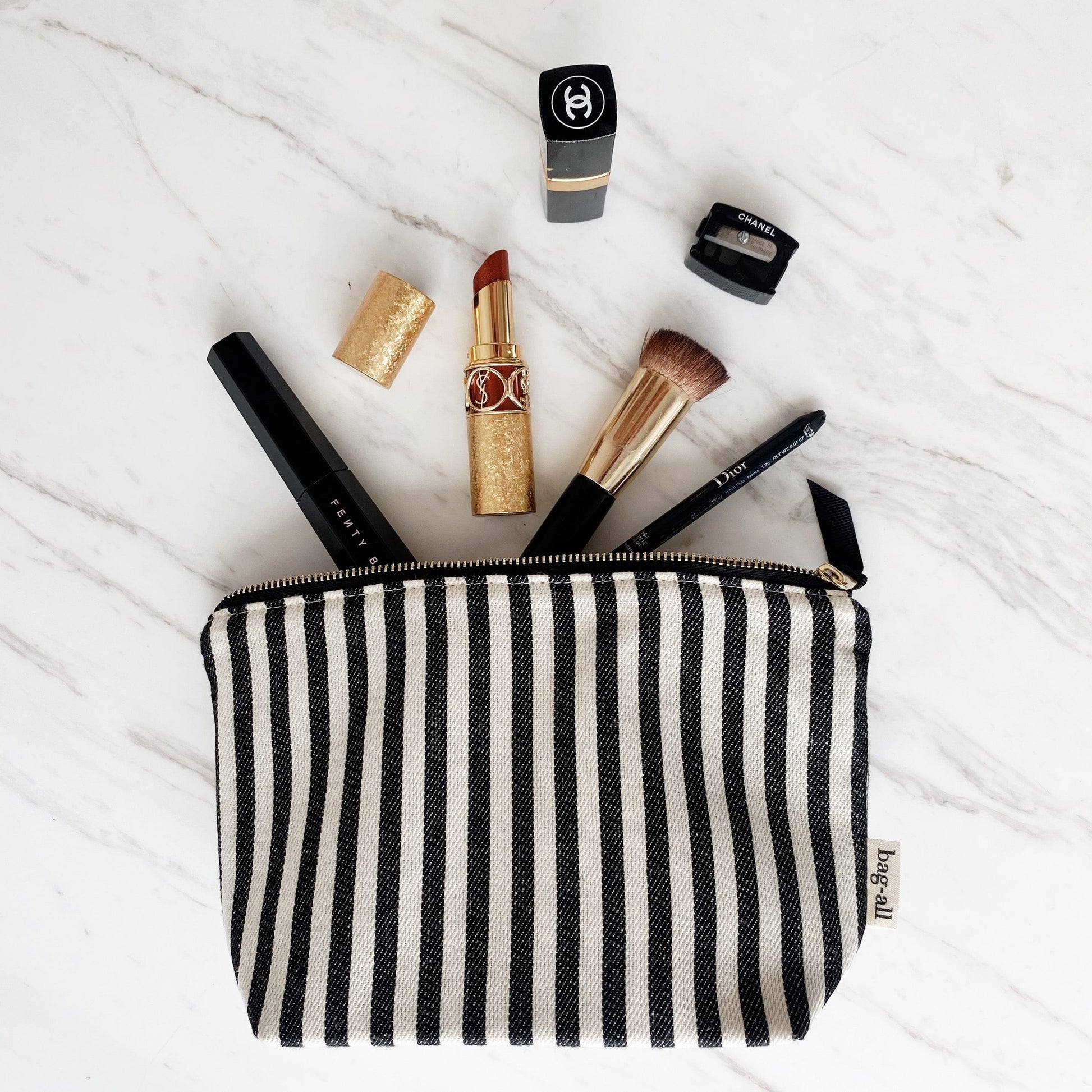 Bag-all Striped Makeup Pouch with gold lipstick, makeup brush, and cosmetics displayed on marble surface, featuring black and white stripes and gold zipper, showcasing practical storage