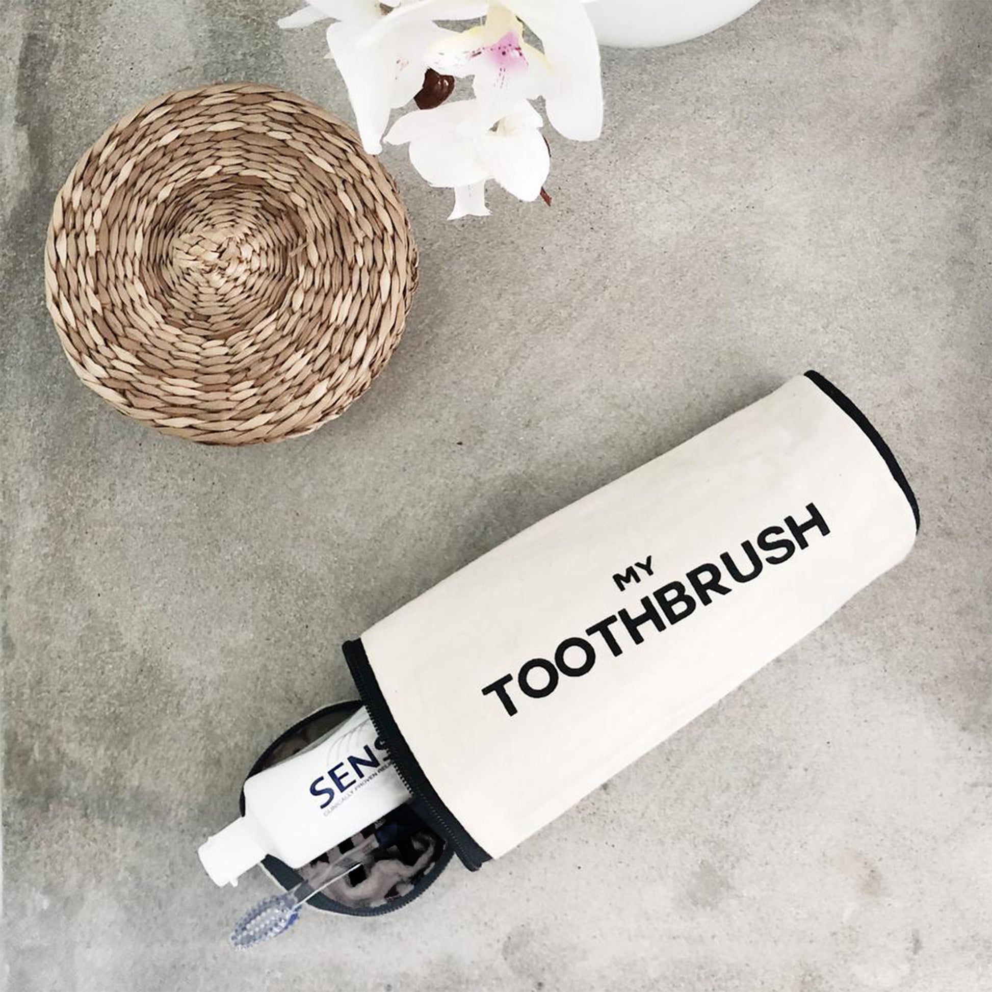 Bag-all cream toothbrush travel case with "My Toothbrush" text shown next to woven coaster and white orchids on concrete surface, minimalist Scandinavian design