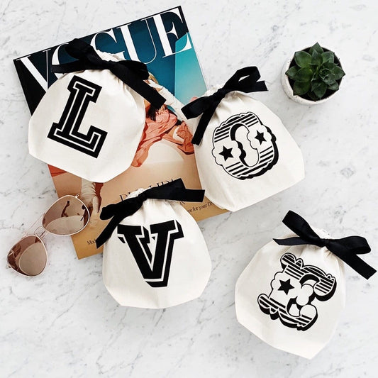 Bag-all Small Letter Bags Mix in cream with black monogram letters and decorative designs, featuring black ribbon ties, displayed on marble surface with Vogue magazine and sunglasses