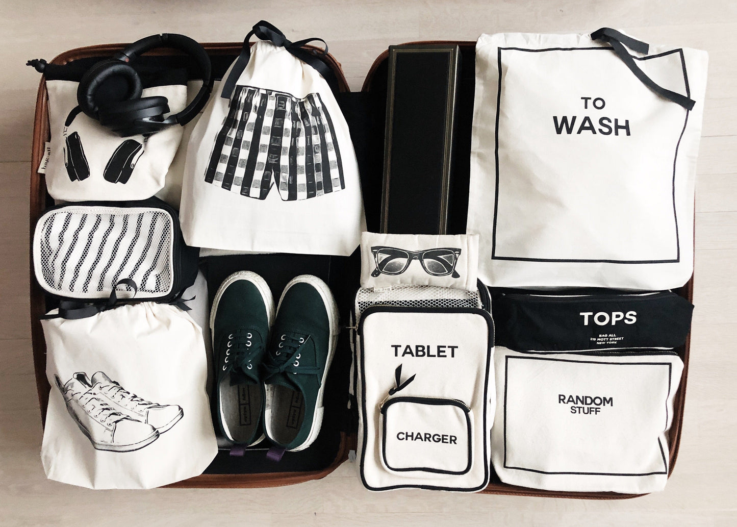 Bag-all White Sneaker Shoe Bag in cream cotton, shown with organized travel essentials including headphones, clothing bags, and tech accessories in monochromatic black and white layout