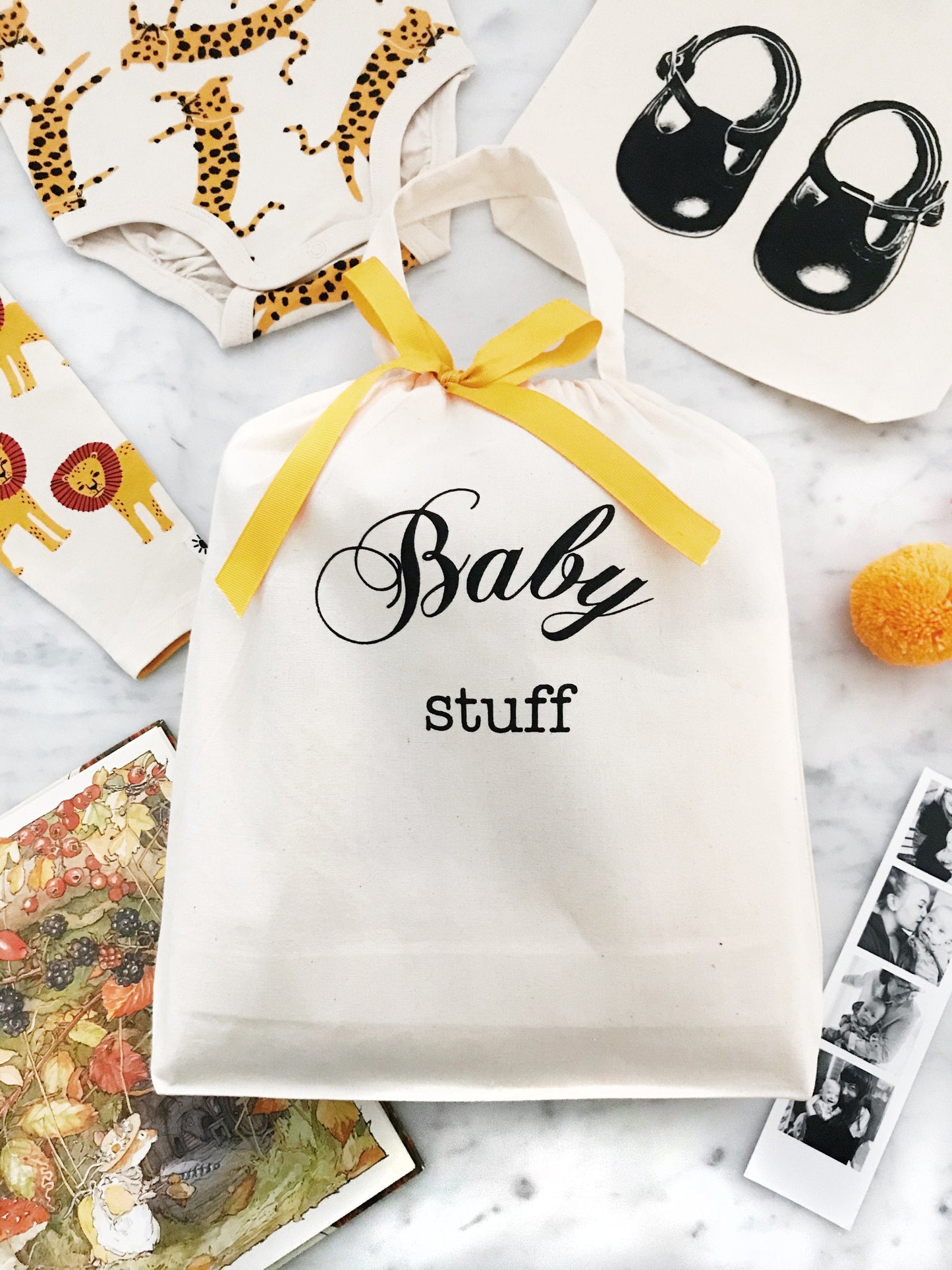 Bag-all Baby Fancy Bag in Cream with yellow ribbon, surrounded by baby essentials like a onesie and illustrated books. Stylish and practical storage