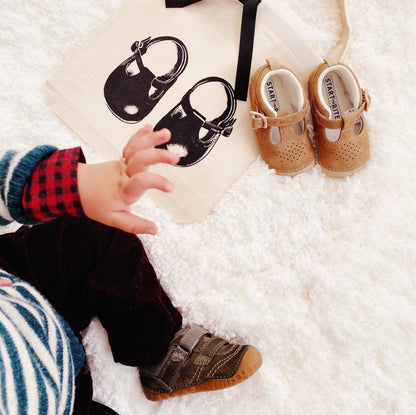 Bag-all Baby Shoe Bag in cream with black bow, pictured with baby shoes and a toddler. Stylish and eco-friendly shoe storage