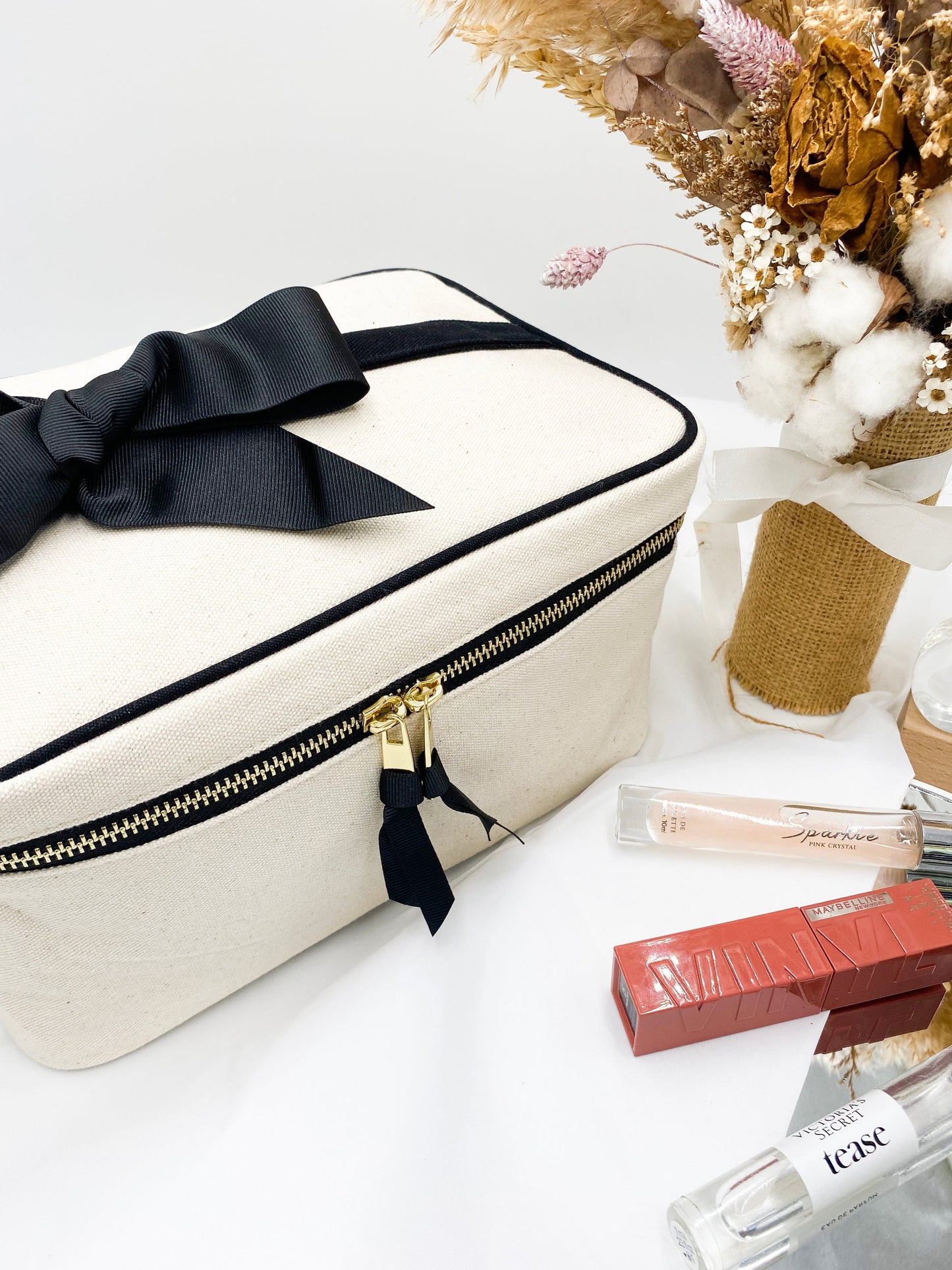 Bag-all Medium Box Makeup & Toiletry Case in cream cotton with black bow detail, gold zipper, and striped interior, styled with beauty products and dried flowers